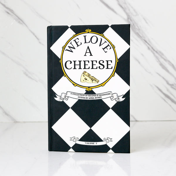 Chef April Burke’s Book titled 'We Love A Cheese' with a black and white checkered design on a white marble background