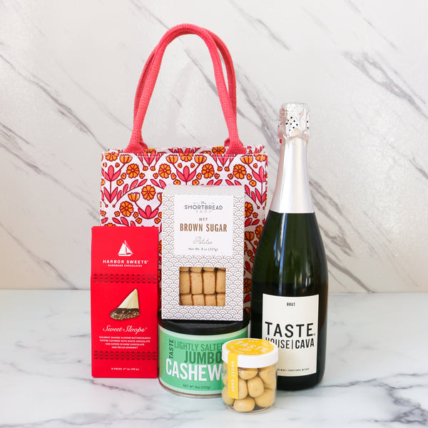 TASTE.online Gift set with a bottle of TASTE House Cava Sparkling wine, cookies, and nuts on a marble surface with a striking pink and yellow Gift Bag