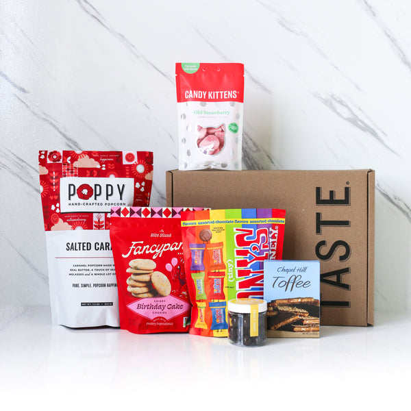 Assorted snack packages in front of a cardboard box with 'Taste' branding on a white background.