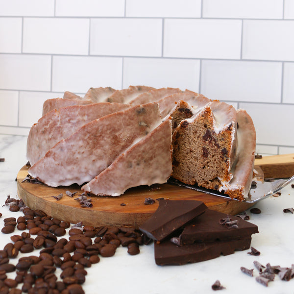 Chocolate chip cake with a slice cut, on a wooden board with chocolate chips and bars on a white tiled background.
