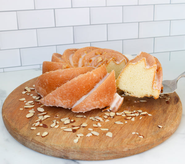 Sliced TASTE bakehouse Amaretto bundt cake with glaze on a wooden board with almonds, against a white tiled wall.