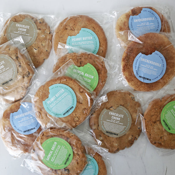 A dozen assorted giant TASTE Bakehouse cookies in individual packaging with different flavors indicated by labels.