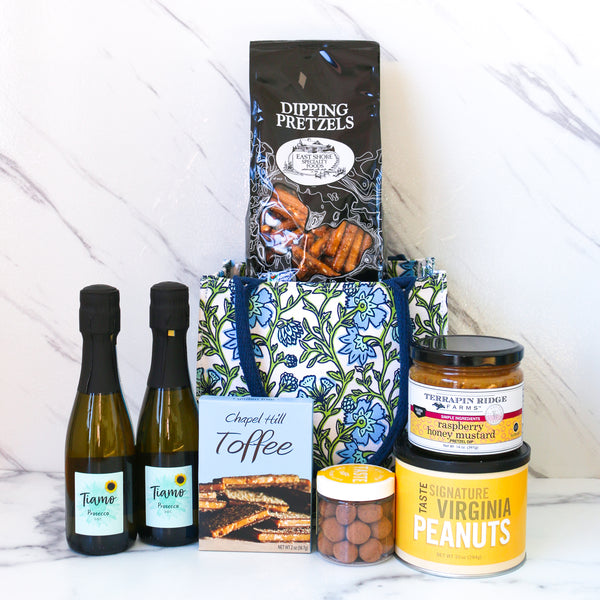 TASTE Gifts A Lil Something. One of TASTE's amazing gifts Assorted snacks and drinks including pretzels, two bottles of prosecco, and peanuts in a decorative bag on a marble background.
