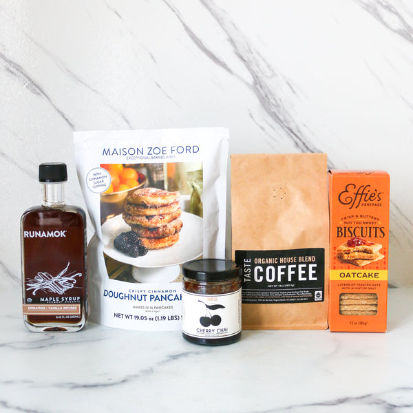 Collection of food products including a bottle of maple syrup, a bag of doughnut pancakes, a jar of cherry jam, a box of coffee, and a box of biscuits on a marble surface.