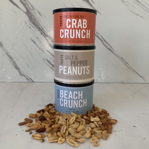 Three cans of TASTE brand snack mixes, arranged from bottom to top with Beach Crunch, Salt & Pepper, and Crab Crunch flavors, surrounded by scattered peanuts and nut shells on a marble surface.