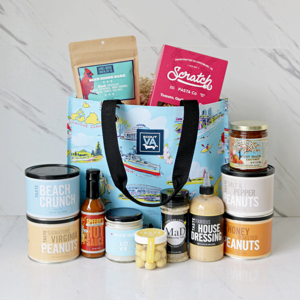 A collection of Virginia-themed food and beverages displayed in front of a reusable SCOUT Virginia bag with a Virginia map pattern. Products include various packaged snacks, bottles of hot sauce and pasta, along with a candle and a magazine.