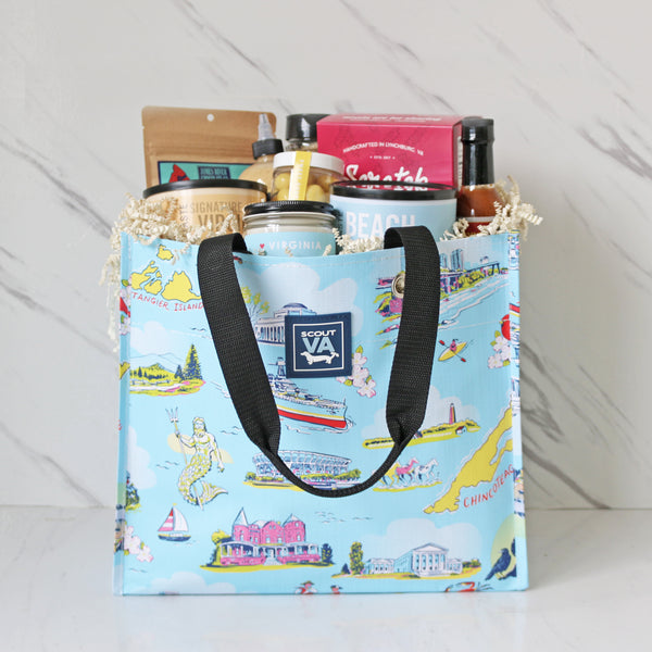 A collection of Virginia-themed food and beverages displayed inside of a reusable SCOUT Virginia bag with a Virginia map pattern. Products include various packaged snacks, bottles of hot sauce and pasta, along with a candle and a magazine.