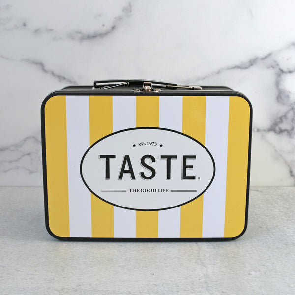 A yellow and white striped metal lunch box with TASTE branding, accompanied by a bottle of dressing, two packs of peanuts, and containers of lemon shortbread bites and celestial sours candies.