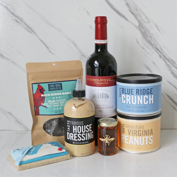 A collection of Virginia-themed food items including a bottle of Barboursville Cabernet Sauvignon, a jar of Virginia Bee Co. Queen Ann Honey, a container of James River Chocolate Blue Ridge Bark, a bottle of TASTE Famous House Dressing, a container of TASTE Blue Ridge Crunch, a bag of TASTE Signature Virginia Peanuts, and a package of TASTE Bakehouse Virginia Cookie.