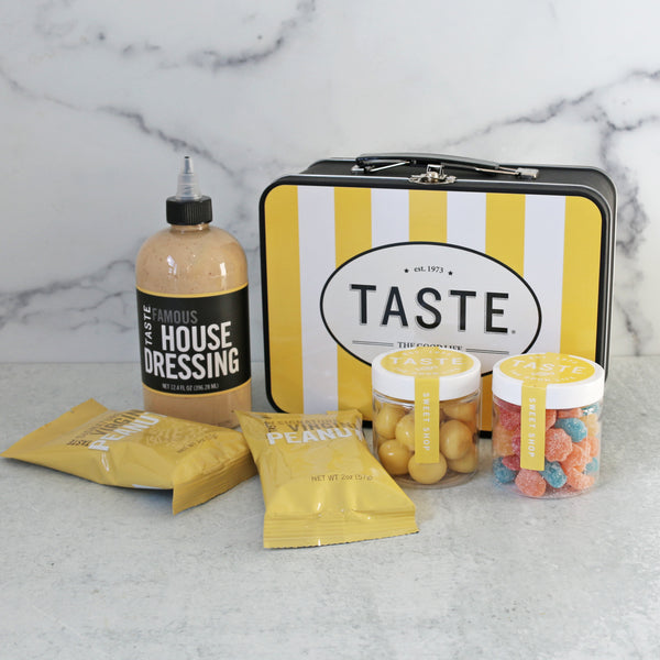 A yellow and white striped metal lunch box with TASTE branding, accompanied by a bottle of dressing, two packs of peanuts, and containers of lemon shortbread bites and celestial sours candies.