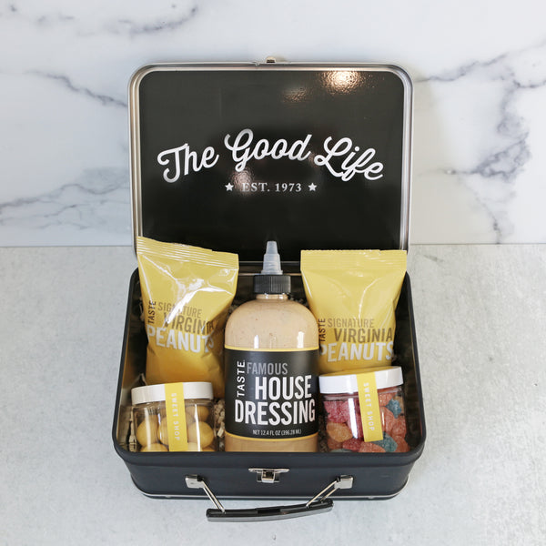 A yellow and white striped metal lunch box with TASTE branding, accompanied by a bottle of dressing, two packs of peanuts, and containers of lemon shortbread bites and celestial sours candies.