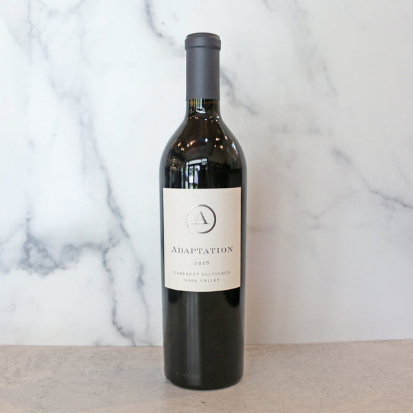 A bottle of 2018 Adaptation Oakville Cabernet Sauvignon wine, standing upright on a marble surface with a plain background.