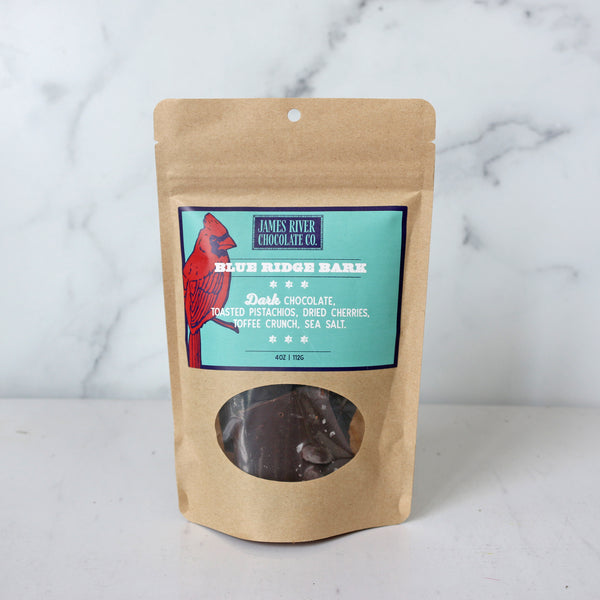 A packaged product labeled 'Blue Ridge Bark' by James River Chocolate Co., visible through a window on a brown paper bag, set against a marble background.