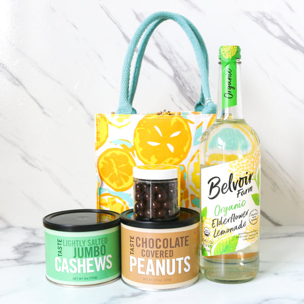 Bottle of Belvoir Farm Organic Elderflower Lemonade, can of chocolate covered peanuts, can of cashews, and a tote bag with lemon design on a white background.