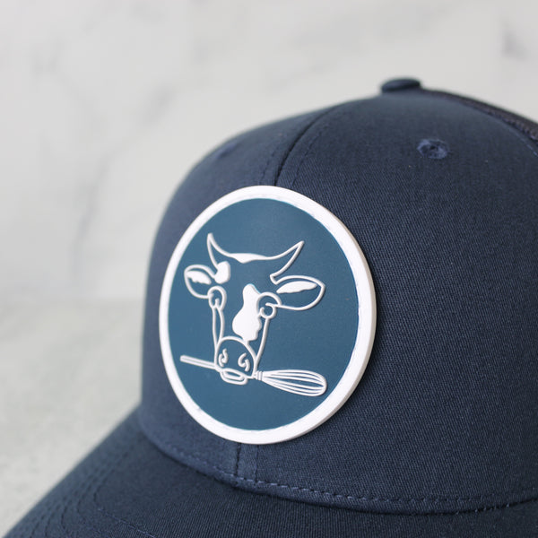 Navy blue trucker hat with the iconic TASTE Bakehouse cow logo on a light gray background featuring a close-up of the logo