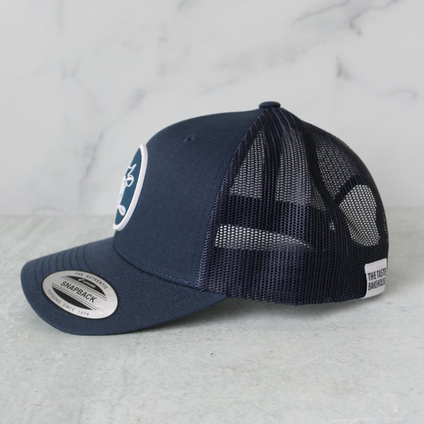 Navy blue trucker hat with the iconic TASTE Bakehouse cow logo on a light gray background featuring a side shot photo