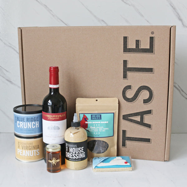 A collection of Virginia-themed food items including a bottle of Barboursville Cabernet Sauvignon, a jar of Virginia Bee Co. Queen Ann Honey, a container of James River Chocolate Blue Ridge Bark, a bottle of TASTE Famous House Dressing, a container of TASTE Blue Ridge Crunch, a bag of TASTE Signature Virginia Peanuts, and a package of TASTE Bakehouse Virginia Cookie.