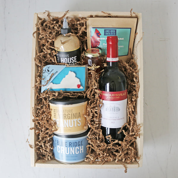 A collection of Virginia-themed food items including a bottle of Barboursville Cabernet Sauvignon, a jar of Virginia Bee Co. Queen Ann Honey, a container of James River Chocolate Blue Ridge Bark, a bottle of TASTE Famous House Dressing, a container of TASTE Blue Ridge Crunch, a bag of TASTE Signature Virginia Peanuts, and a package of TASTE Bakehouse Virginia Cookie.