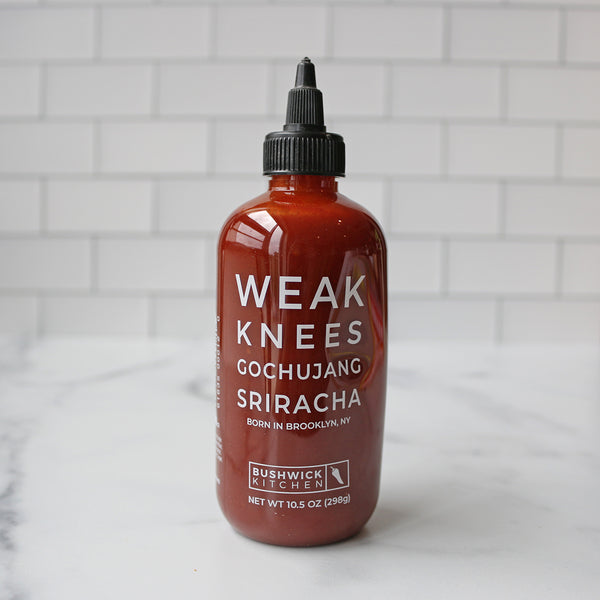 A bottle of Bushwick Kitchen Weak Knees Gochujang Sriracha hot sauce on a marble surface with white tile background.