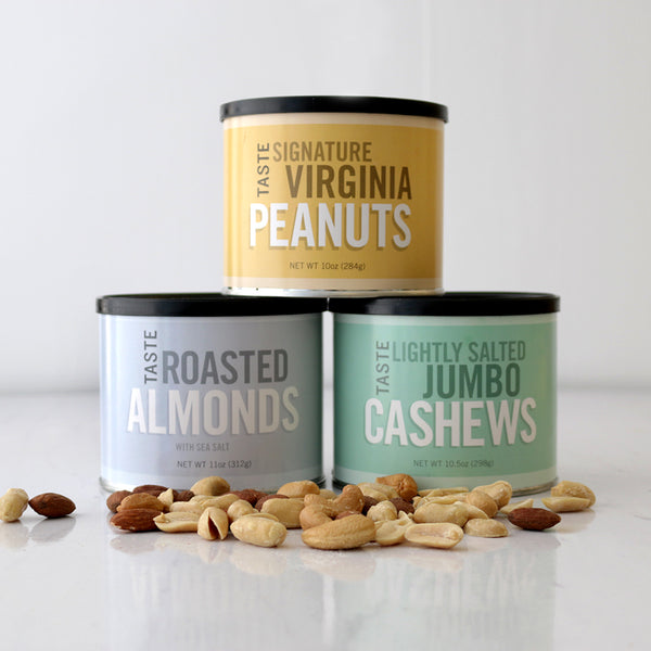 Three tin containers of nuts arranged on a white surface, with roasted almonds, virginia peanuts, and lightly salted jumbo cashews spilling out in front of the containers.