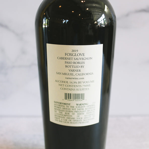 the back of a bottle of Foxglove Cabernet Sauvignon with a label that includes the wine variety and the brand name. The bottle is upright and has a red cap, set against a marble background.