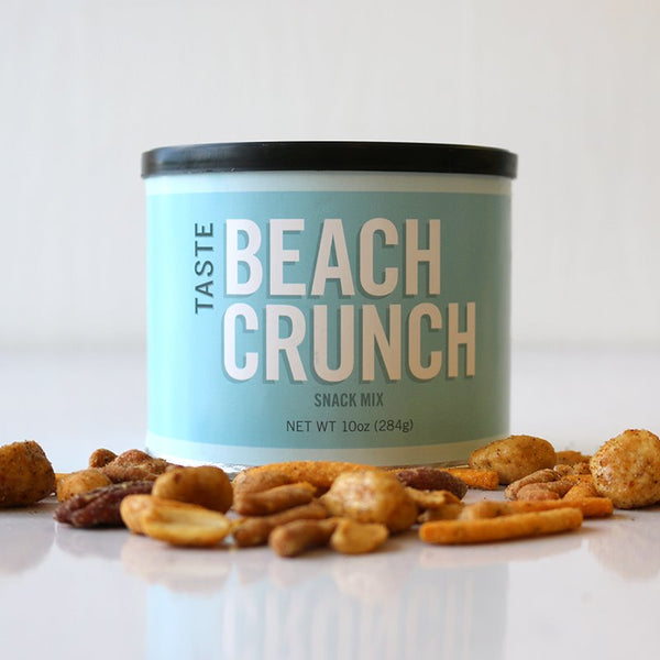 A can of TASTE Beach Crunch snack mix with various nuts spilled in front of it on a white surface.