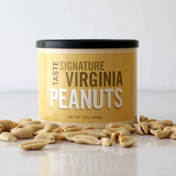 A yellow cylindrical container labeled 'TASTE Signature Virginia PEANUTS' with a black lid, surrounded by lightly salted peanuts scattered on the surface.