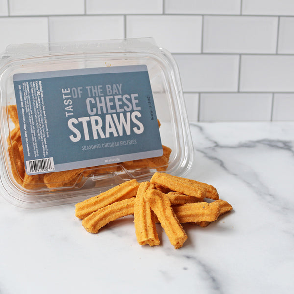 A resealable plastic container of TASTE of the Bay Cheese Straws on a marble surface displaying some cheese straws.