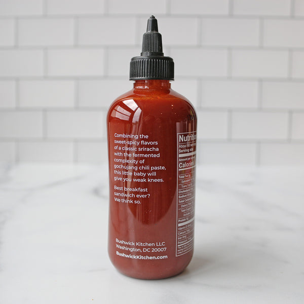 A bottle of Bushwick Kitchen Weak Knees Gochujang Sriracha hot sauce on a marble surface with white tile background.