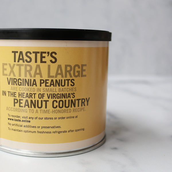 A yellow cylindrical container labeled 'TASTE Signature Virginia PEANUTS' with a black lid, surrounded by lightly salted peanuts scattered on the surface.