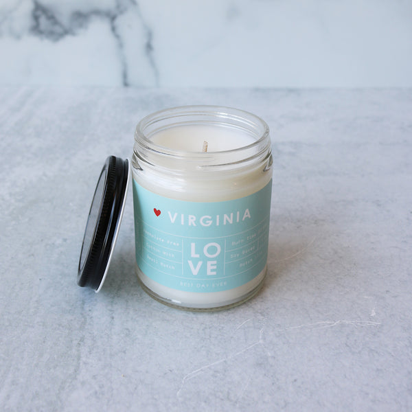 A jar candle with a white label that reads 'VIRGINIA LOVE' on a blue and green striped background, placed on a marble surface.